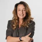 Smiling woman with wavy light brown hair in a gray blazer, arms crossed, wearing a watch, against a plain light background.
