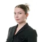 Portrait of a young woman with light skin, wearing a black blazer with a fabric rose detail, her hair in a bun, against a white background.