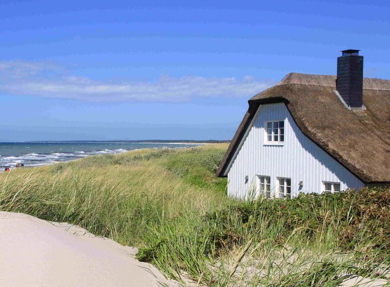 A white cottage with a thatched roof sits beside tall grass dunes overlooking a sandy beach and ocean under a clear blue sky.