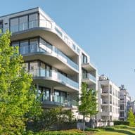 Modern white apartment building with curved glass balconies framed by green trees under a clear blue sky.