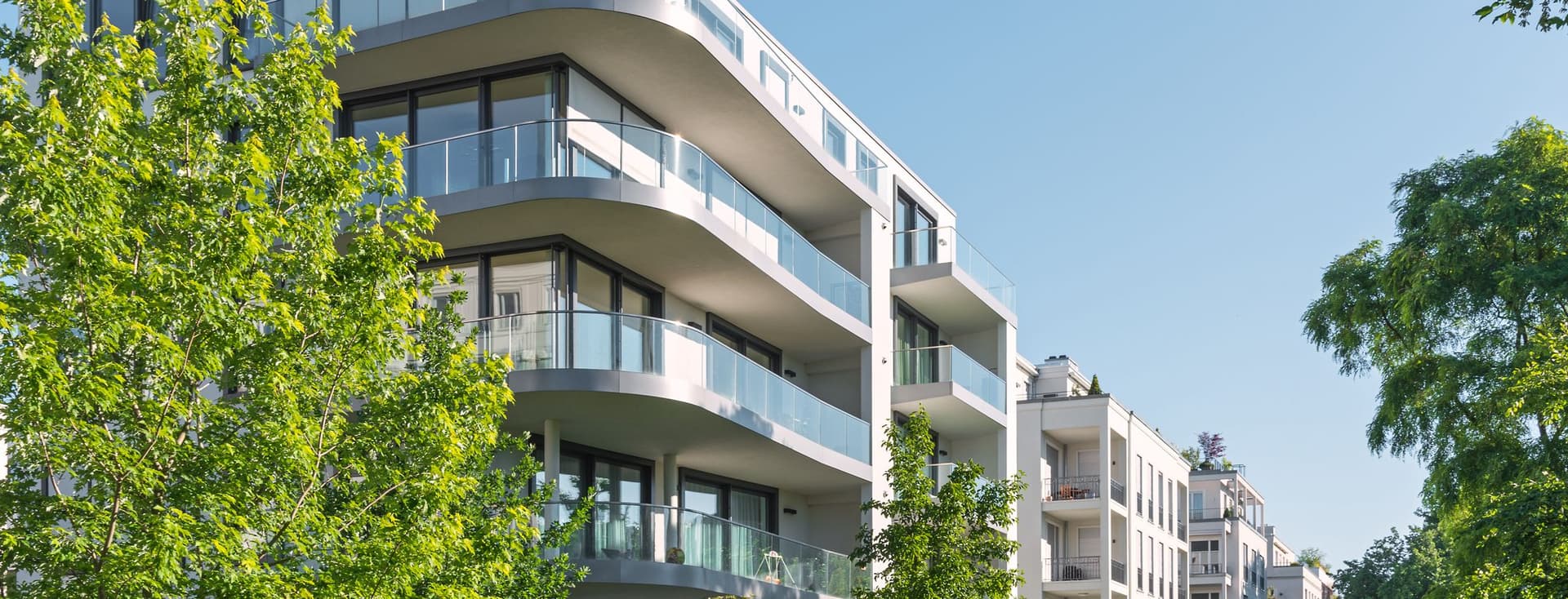Modern white apartment building with curved glass balconies framed by green trees under a clear blue sky.