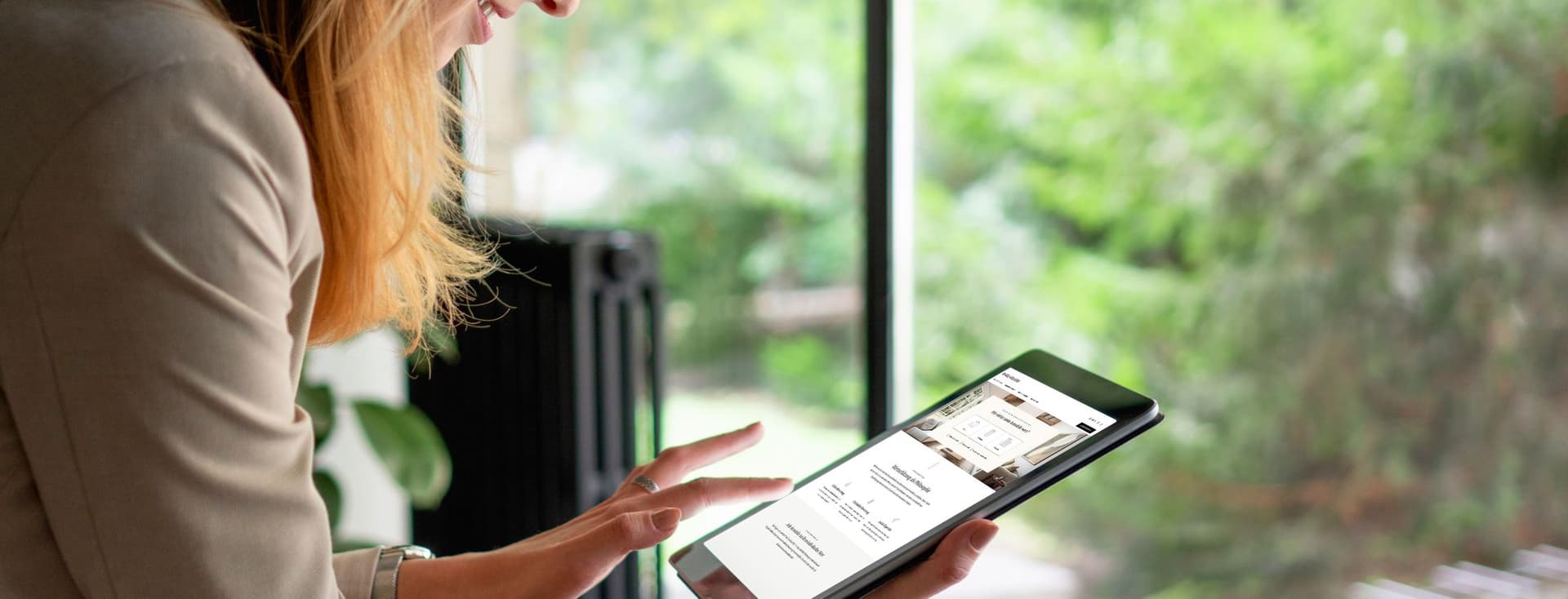 A person in a beige blazer sitting by a window while reviewing digital content on a tablet device in a bright, naturally lit setting