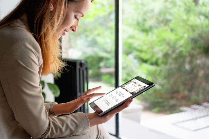 A person in a beige blazer sitting by a window while reviewing digital content on a tablet device in a bright, naturally lit setting