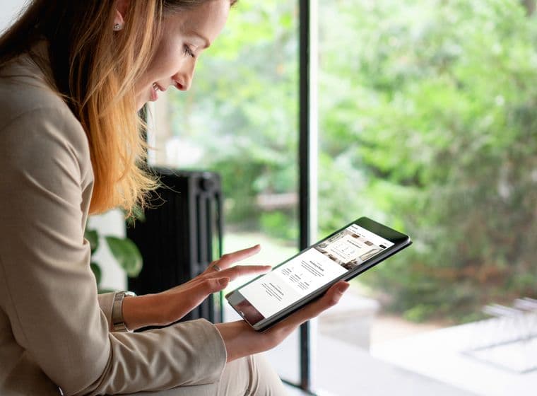 A person in a beige blazer sitting by a window while reviewing digital content on a tablet device in a bright, naturally lit setting