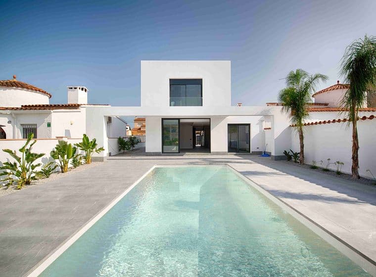 Vista d'una casa moderna blanca amb piscina a l'entrada, jardí, palmeres al voltant i cels blaus.