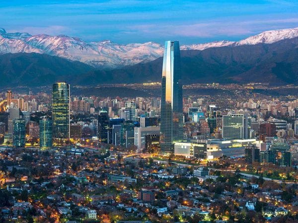 Edificio moderno con fachada de formas geométricas y una gran entrada de cristal, rodeado de árboles y arbustos cuidadosamente ajardinados bajo un cielo azul.