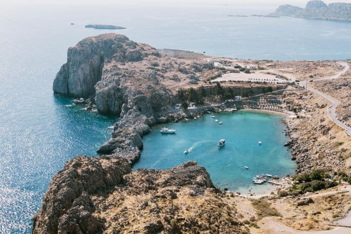 Aerial view of a rocky coastline with a small bay, clear turquoise water, boats, and a winding road on a sunny day.