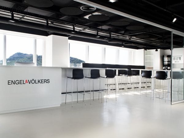 Bright, modern office interior with white counters labeled Engel & Völkers, exposed ceiling ducts, seating area, and large windows with a city view.