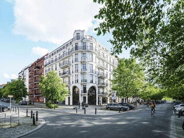 Elegant white corner building with historic architecture, featuring arched ground floor entrances, decorative balconies, and a distinctive rounded corner tower, set on a tree-lined street with parked cars and cyclists