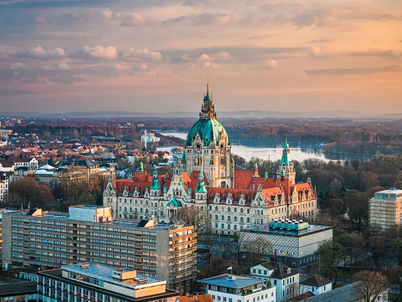 Aerial sunset view of a Hannover; ornate central building with green copper dome, red-tiled roofs, and a winding river in the distance.