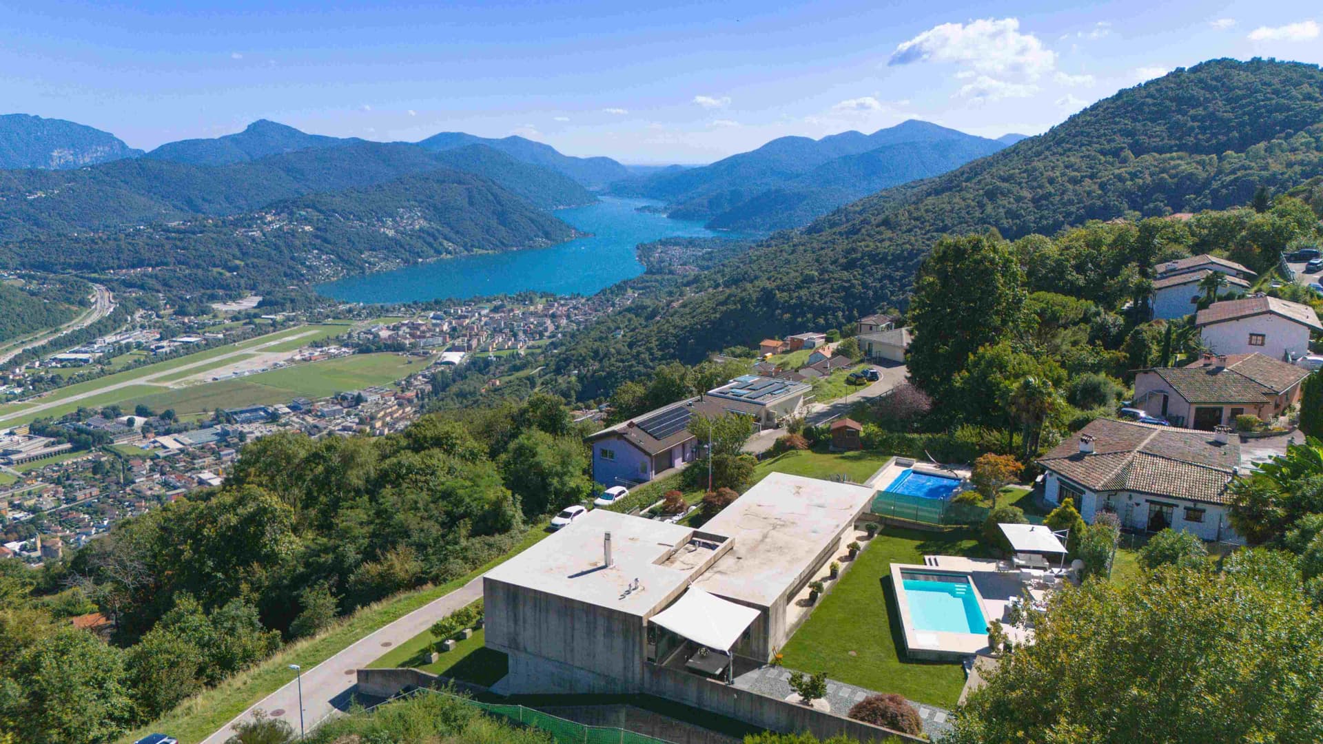 Aerial view of a turquoise lake in a mountain valley, with forested hills and hillside homes with swimming pools in foreground.