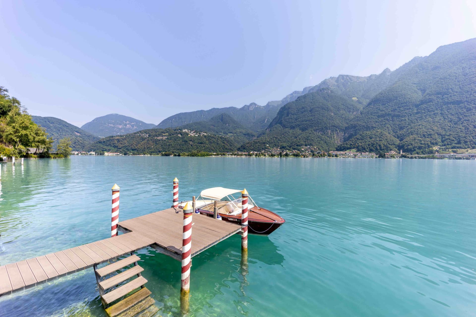 Wooden dock with striped poles and a small boat on a turquoise lake, forested mountains and clear sky in the background.
