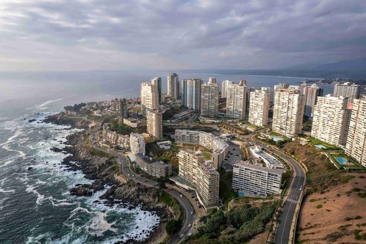 Vista aérea de una ciudad costera con edificios blancos de gran altura, carreteras sinuosas y costa rocosa bajo un cielo nublado.