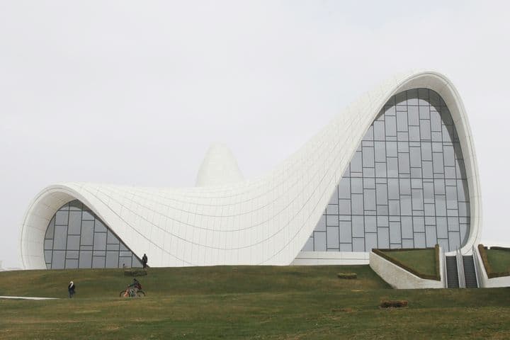 Modern architectural building featuring dramatic curved white waves in its design, characteristic of Zaha Hadid's style. The structure's flowing lines create an undulating roof against a grey sky, with small figures visible on the grassy slope in front for scale.