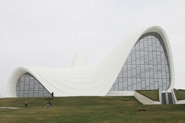 Modern architectural building featuring dramatic curved white waves in its design, characteristic of Zaha Hadid's style. The structure's flowing lines create an undulating roof against a grey sky, with small figures visible on the grassy slope in front for scale.
