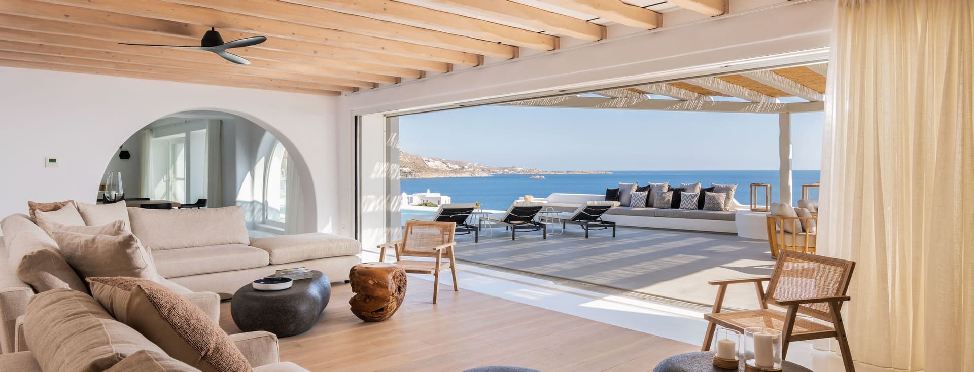 Bright living room with beige sofas, wood tables, and open doors to a patio with ocean views. Ceiling has exposed wood beams.