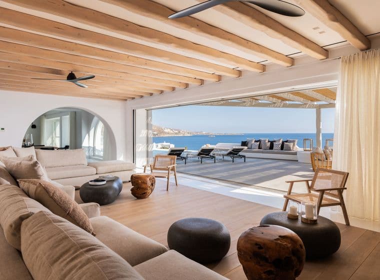 Bright living room with beige sofas, wood tables, and open doors to a patio with ocean views. Ceiling has exposed wood beams.