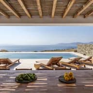 Open veranda with exposed wooden beams framing a calm sea and distant mountains; poolside loungers and bowls of lemons and limes.