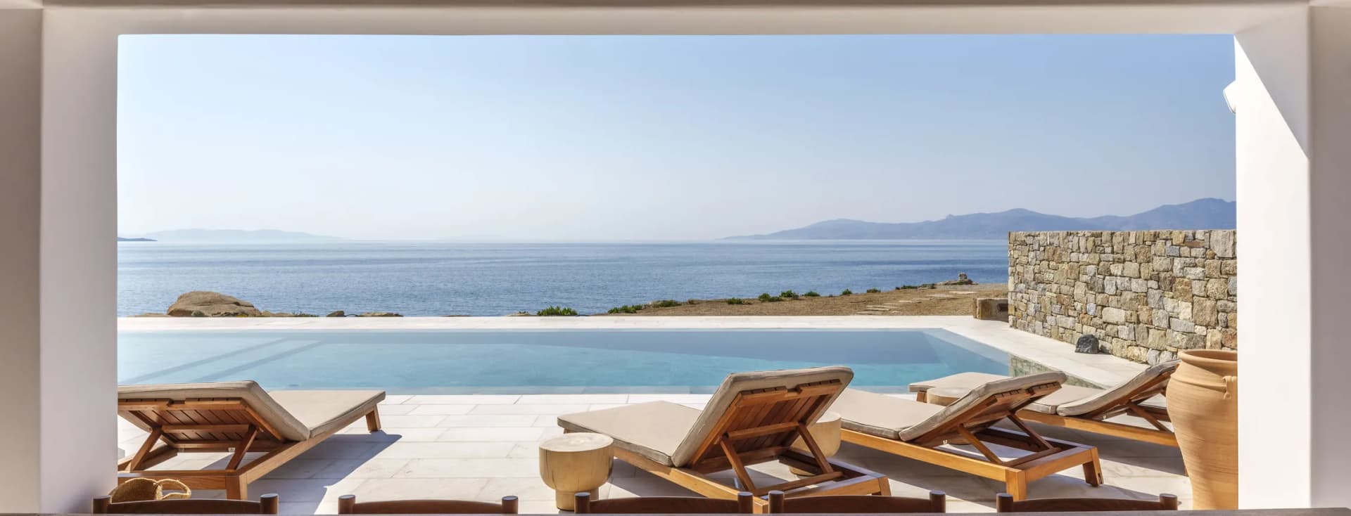 Open veranda with exposed wooden beams framing a calm sea and distant mountains; poolside loungers and bowls of lemons and limes.