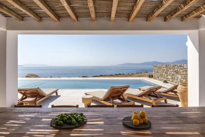 Open veranda with exposed wooden beams framing a calm sea and distant mountains; poolside loungers and bowls of lemons and limes.