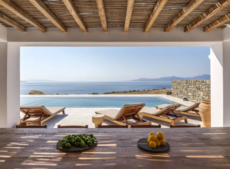 Open veranda with exposed wooden beams framing a calm sea and distant mountains; poolside loungers and bowls of lemons and limes.