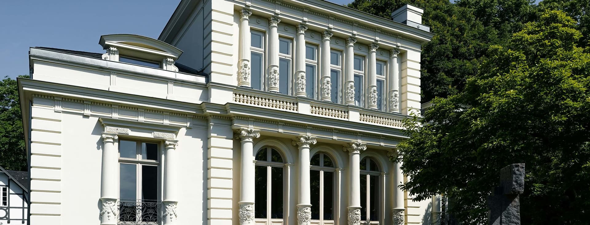 White neoclassical building with tall decorative columns, arched windows, and balconies, beside green trees under a clear blue sky.