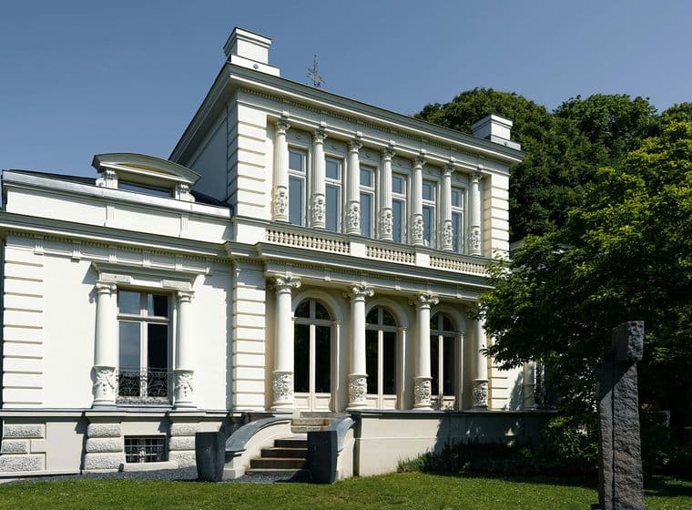 White neoclassical building with tall decorative columns, arched windows, and balconies, beside green trees under a clear blue sky.