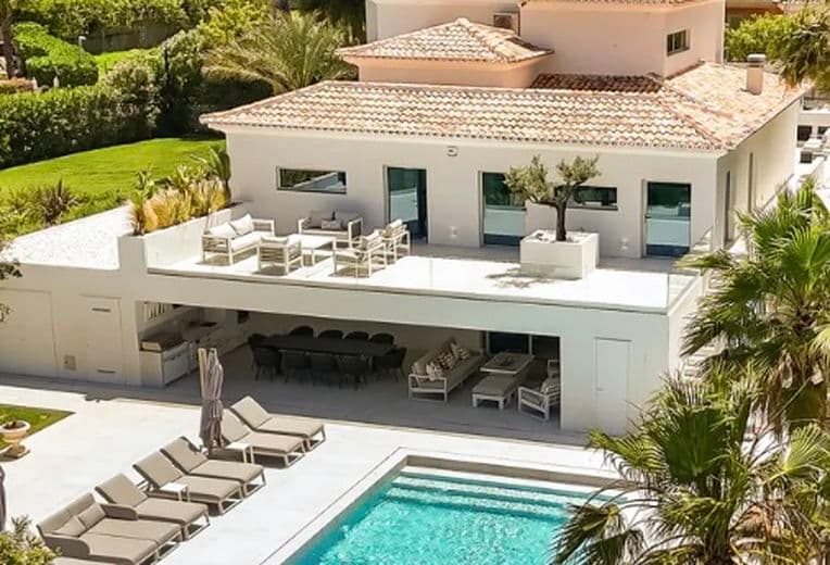 Modern white villa with terracotta roof, glass doors, upper deck seating, and a rectangular blue pool flanked by lounge chairs and palm trees.