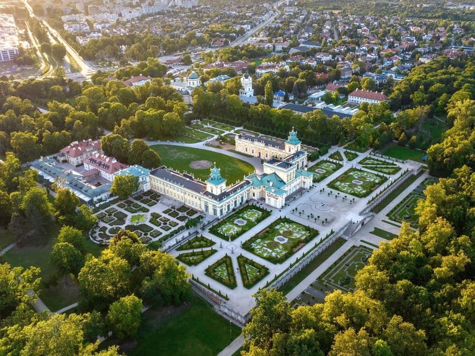 Aerial view of a large, historic palace surrounded by gardens and trees, with a cityscape and sunset in the background.