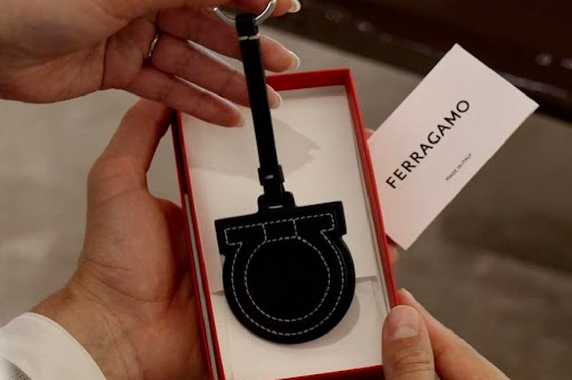 Hands holding an open red Ferragamo box revealing a black leather keychain with white stitching and a Ferragamo card.