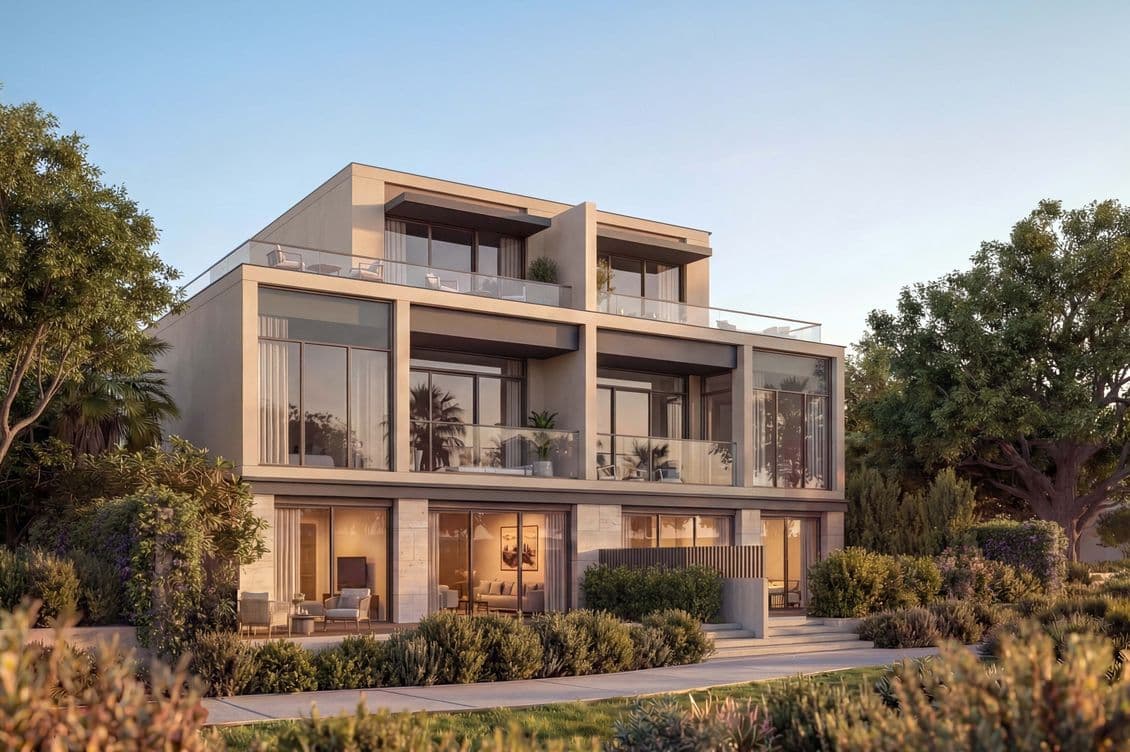 Modern three-story house with large windows and balconies, surrounded by lush greenery and trees, under a clear sky.