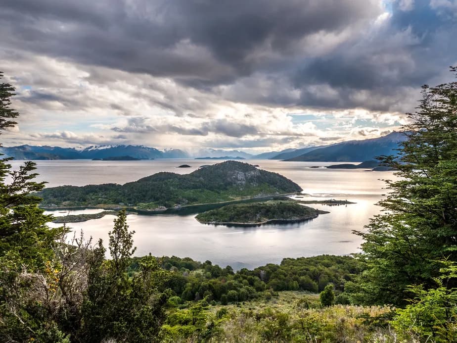 A picturesque view of a lake with small islands, surrounded by lush greenery and mountains beneath a dramatic cloudy sky.