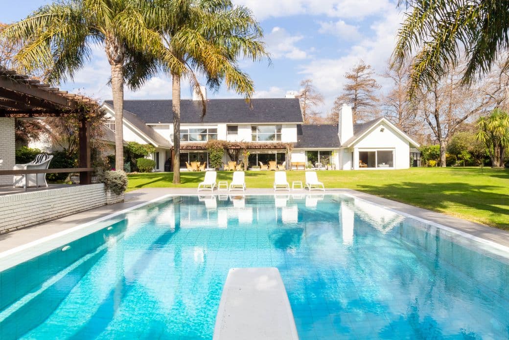 Large modern house with palm trees, green lawn and reflecting blue swimming pool with diving board and lounge chairs.