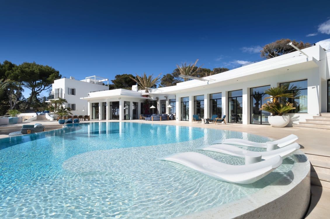 Luxurious villa with large glass windows, a curving infinity pool, white lounge chairs, and lush greenery under a clear blue sky.
