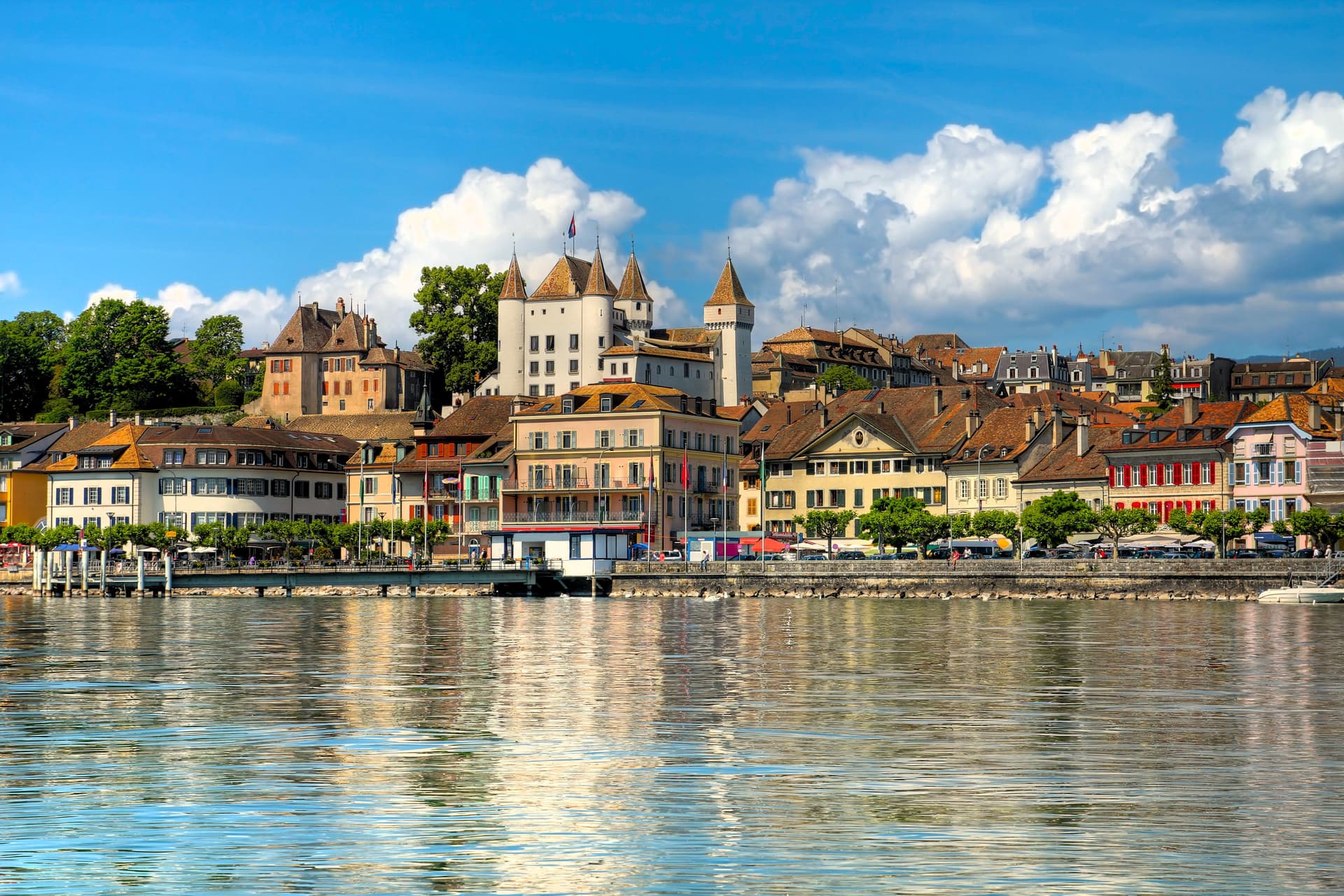 Lakeside European town with castle towers, pastel houses, and a waterfront promenade beneath a bright blue sky.