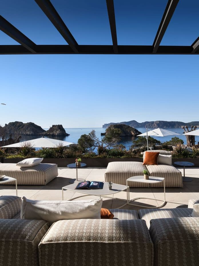A luxurious outdoor terrace with comfortable modern lounge seating and sunbeds under a dark pergola, offering a scenic view of a calm sea bay, rocky islands, and a bright, sunny blue sky.