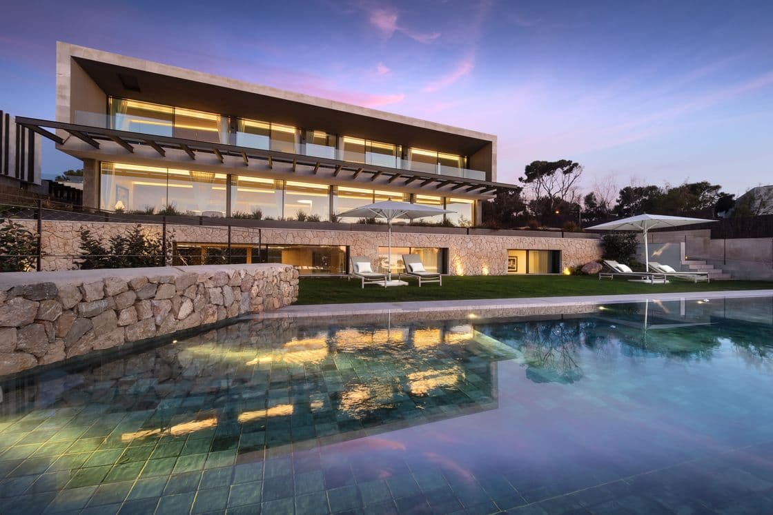 Modern villa with large glass windows, warm lighting, and a stone facade, set against a sunset sky. A pool reflects the evening glow.