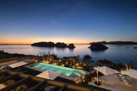Luxury seaside villa at sunset with pool, seating area, and views of islands under a starry sky.