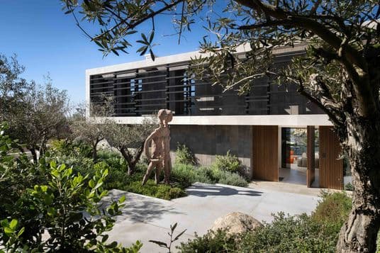 Modern building with a large stone sculpture and lush garden, framed by trees under a clear blue sky.