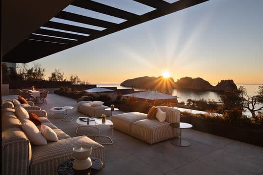 Luxurious terrace overlooking the ocean at sunset, featuring plush seating, tables, and a stunning view of the sun setting behind rocky islands.