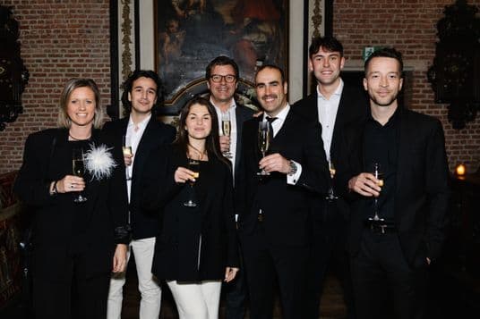 Group of seven professionals in black suits and dresses, raising champagne flutes in a brick-walled room with a large classical painting behind them.