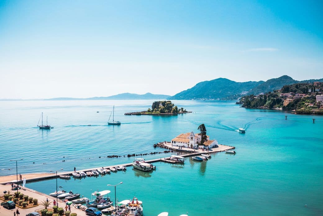 Scenic view of a small, white chapel on a coastal pier in Corfu, Greece, with boats docked around it. The clear turquoise sea surrounds the pier, and mountainous islands are visible in the background under a clear blue sky.