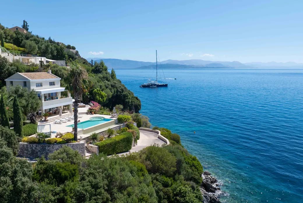 Coastal hillside villa with white terrace, swimming pool, and lush greenery overlooking blue sea; distant sailboat and mountains on the horizon.