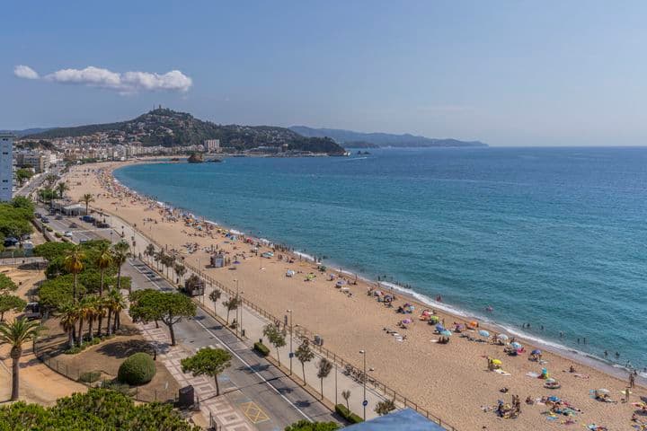 Malerischer Blick auf einen Sandstrand mit türkisfarbenem Wasser, sonnenbadende Menschen und eine Stadt im Hintergrund.