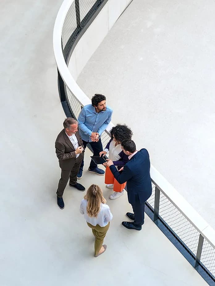 Top-down view of six professionals in business attire standing on a curved walkway, gathered around a tablet.