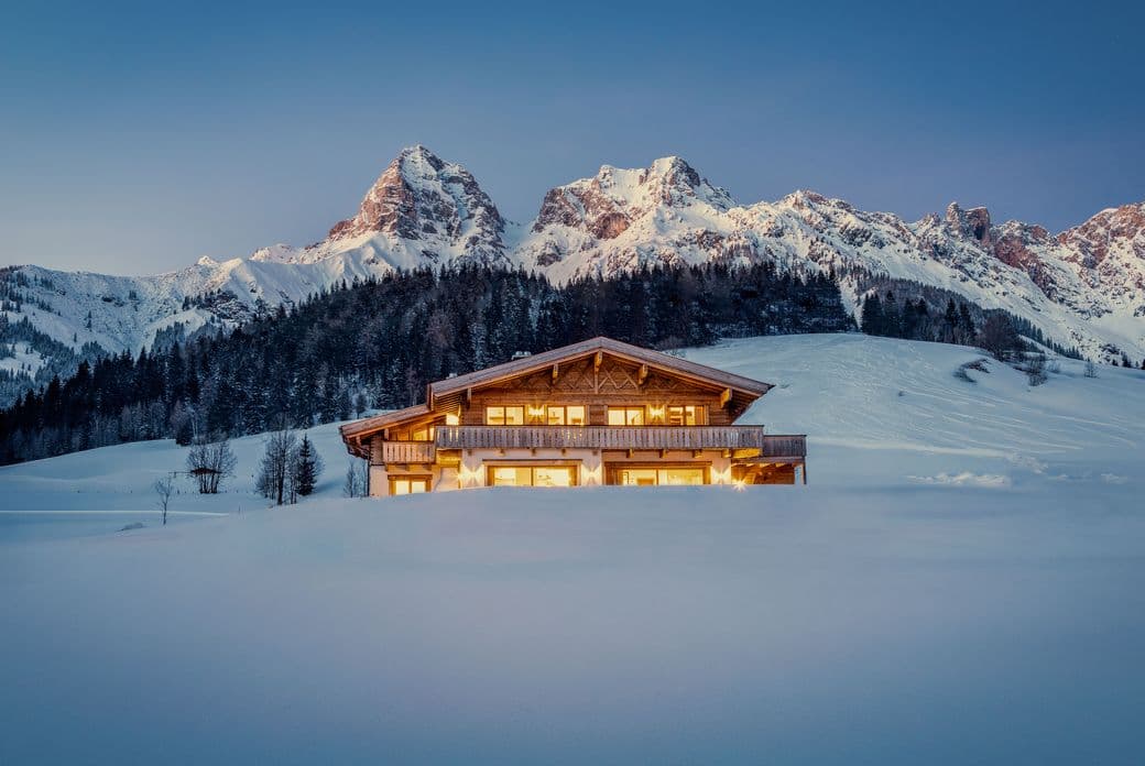 A cozy cabin with glowing windows in a snowy landscape, set against majestic, snow-capped mountains under a clear evening sky.