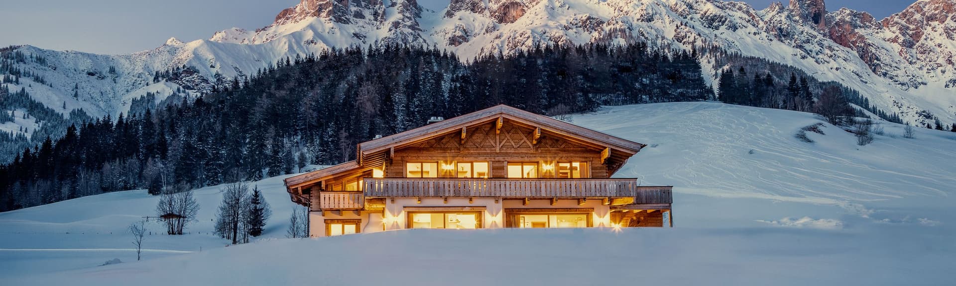 A cozy cabin with glowing windows in a snowy landscape, set against majestic, snow-capped mountains under a clear evening sky.