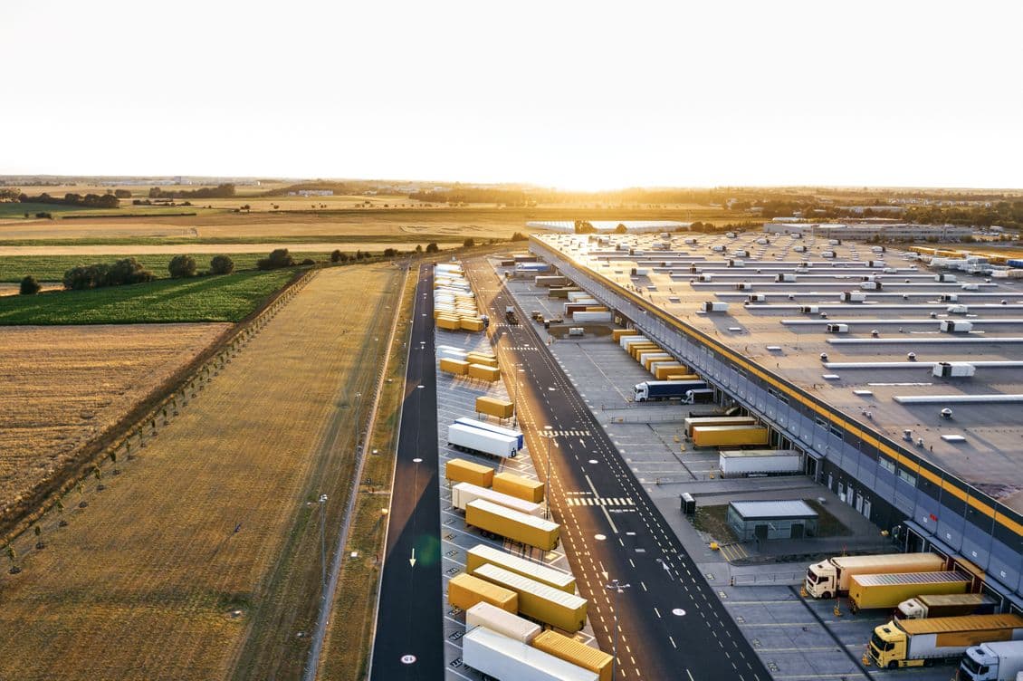 Aerial drone view of a large logistics facility at sunset, showing loading docks with parked trucks, extensive warehouse buildings, and surrounding agricultural fields, photographed from above