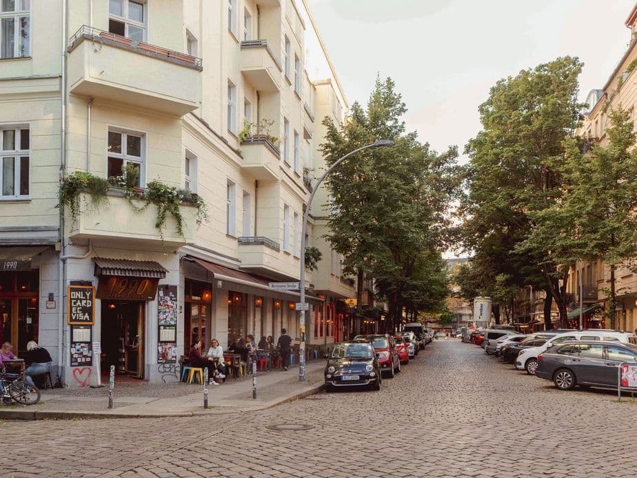 Street scene with a corner cafe, people seated outside, lined with parked cars and bicycles, surrounded by trees and multi-story buildings.