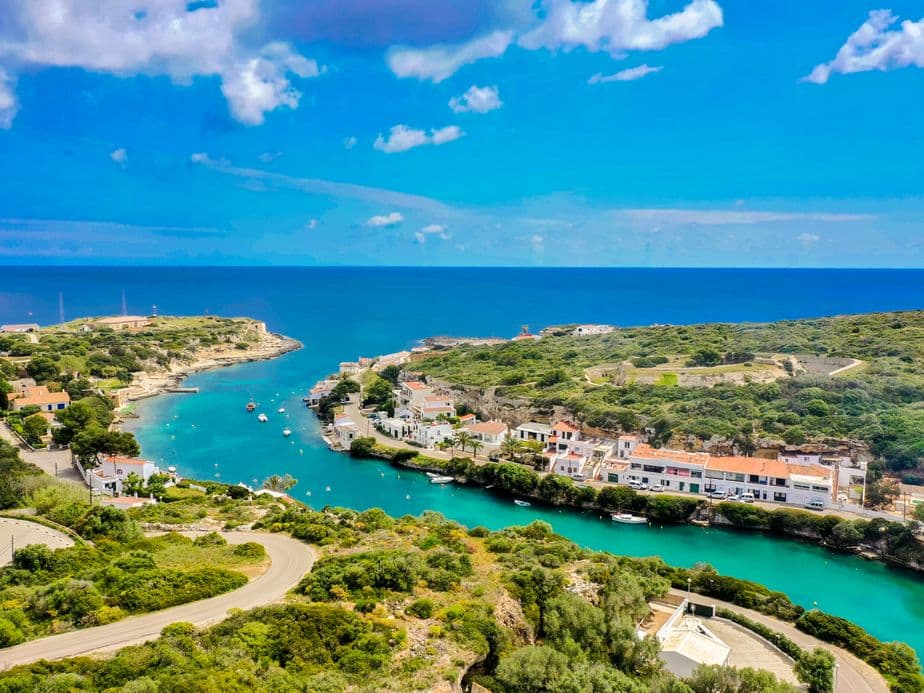 Vista aèria d’una badia tranquil·la a Menorca amb aigües turqueses, petites embarcacions i cases tradicionals a banda i banda, envoltada de turons verds i amb obertura cap al mar Mediterrani.